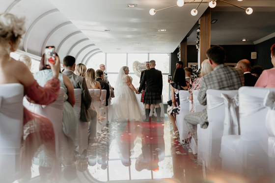 Bride and groom stand at altar holding hands during ceremony, while guests look on from white chairs.